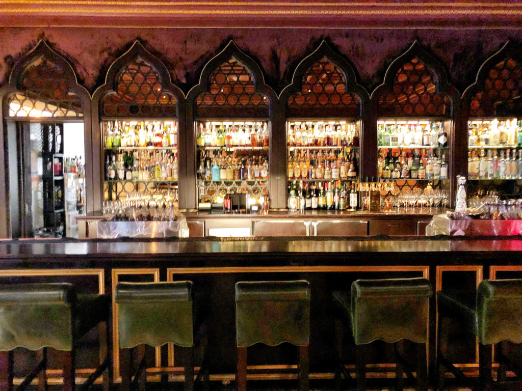 New traditional interior design style - Ornate bar with illuminated liquor display shelves and emerald velvet barstools at the Nomad Hotel, Los Angeles.