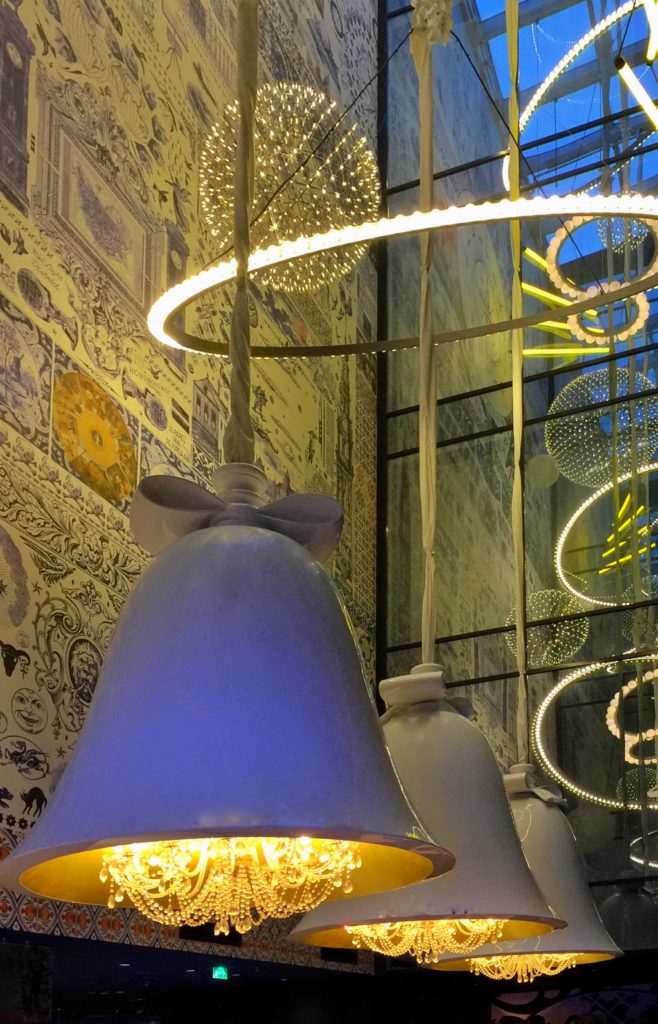 Three hanging bell chandeliers next to astronomy wallpaper in the atrium at the Andaz hotel in Amsterdam express a whimsical interior design style.