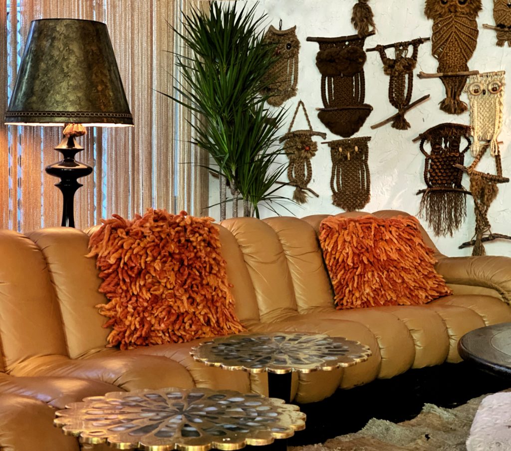 Curved ochre sofa, furry orange throw pilows, a large floor lamp, and some macrame owls create a cozy Boho interior design style.