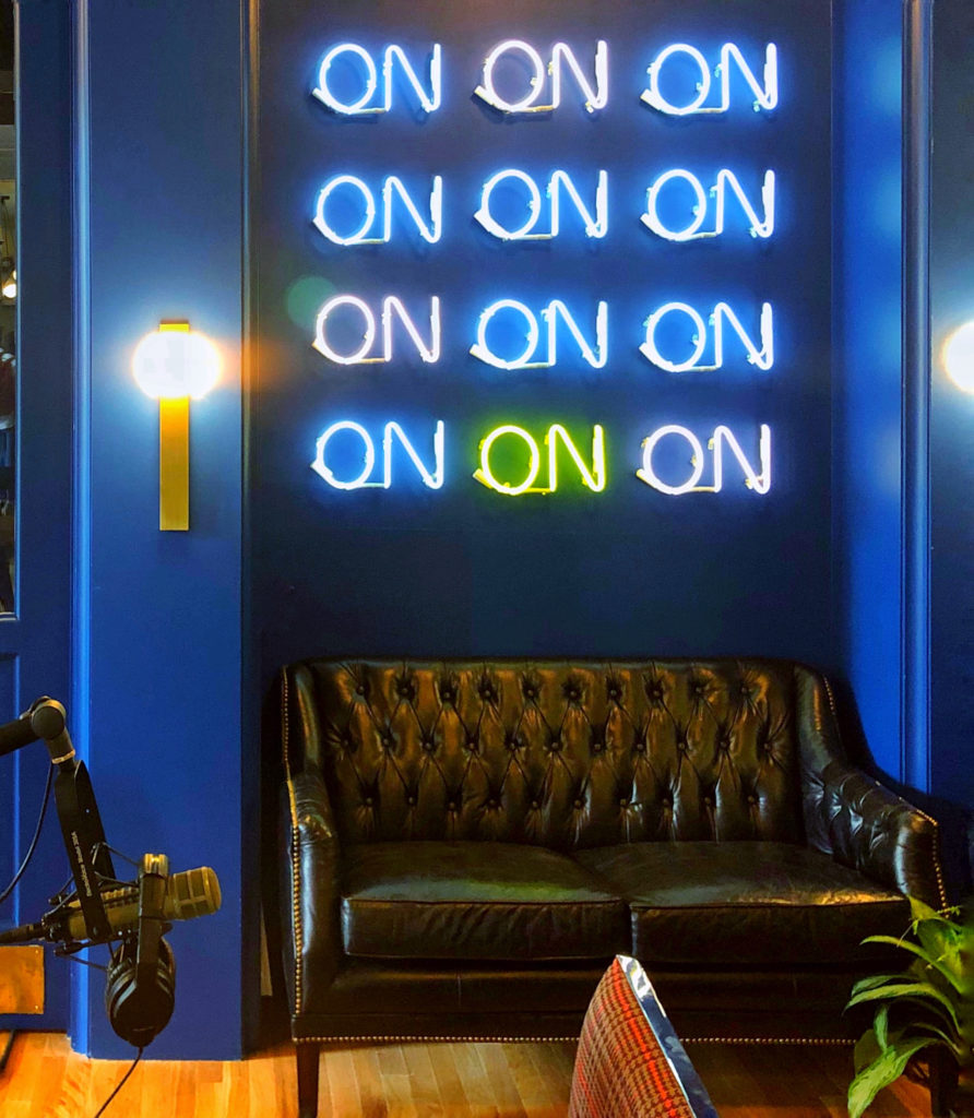Podcast studio with blue walls and neon signage with repeated "ON" letters with a leather sofa and a microphone.