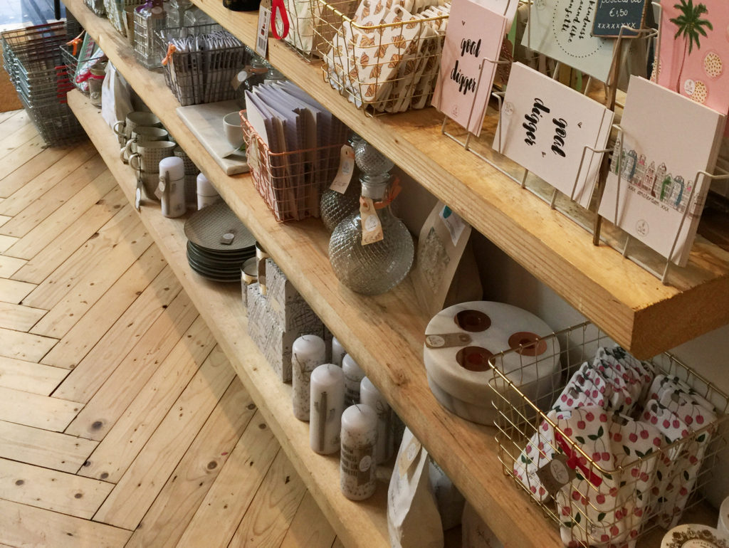 Retail merchandise at Pluk Amsterdam display including cards, candles, and home goods displayed on wood plank shelves and wire metal baskets.