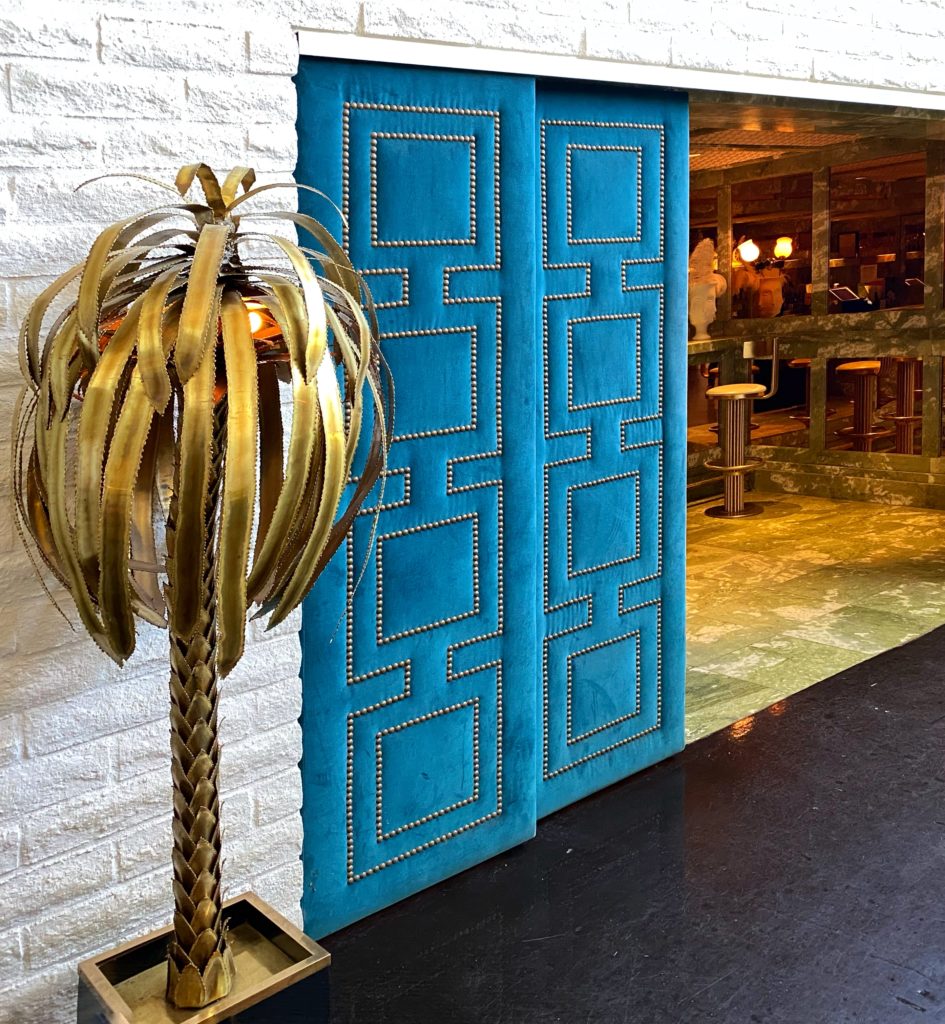 Turquoise velvet doors with nailhead trim detail flanked by two brass palm tree sculptures at the entrance to Mini Bar at the Parker Palm Springs Hotel.