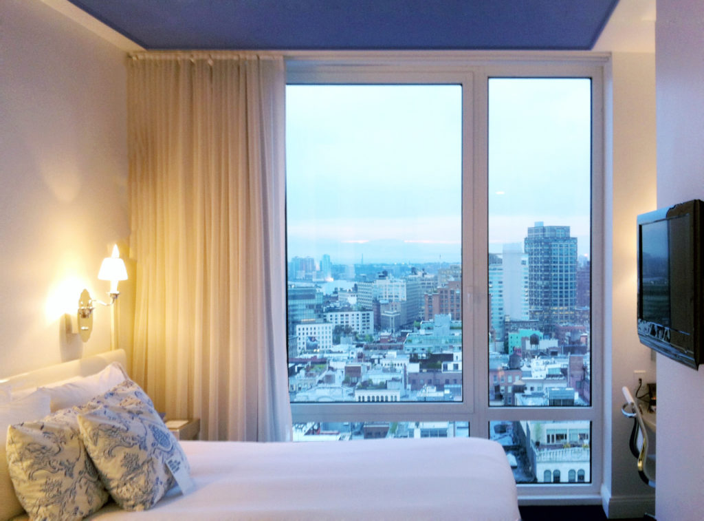Bedroom with blue ceiling and urban skyline outside large picture window.