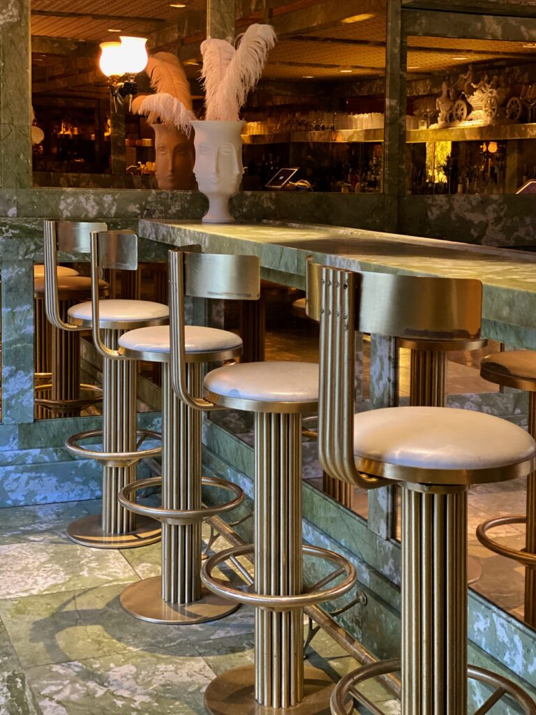 Bohemian decorating at The Parker Palm Springs hotel bar featuring Jonathan Adler’s green marble counter, gold Art Deco bar stools, and sculptural vases with ostrich feathers in a glamorous mid-century desert setting.