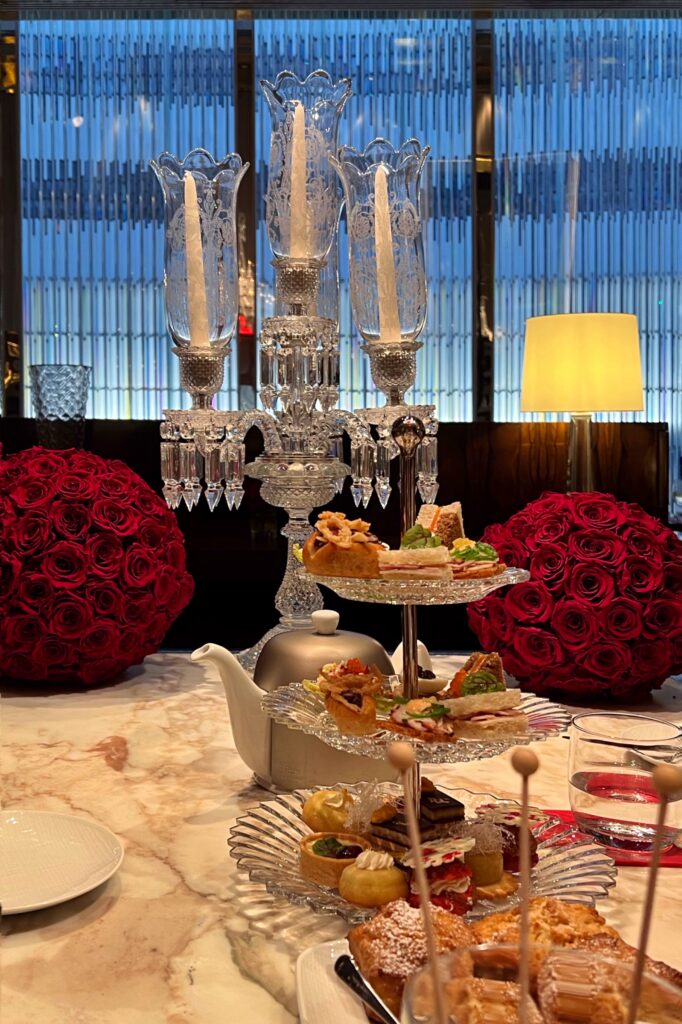Afternoon tea at Baccarat Hotel New York with crystal candelabra, red rose centerpieces, and tiered trays of French pastries and savory bites — a luxury travel and interior design experience in Manhattan.
