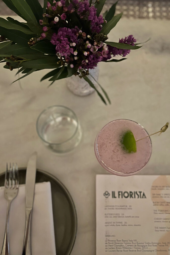 A lavender colored cocktail sits on the marble countertop next to some purple flowers at Il Fiorista restaurant in New York City.