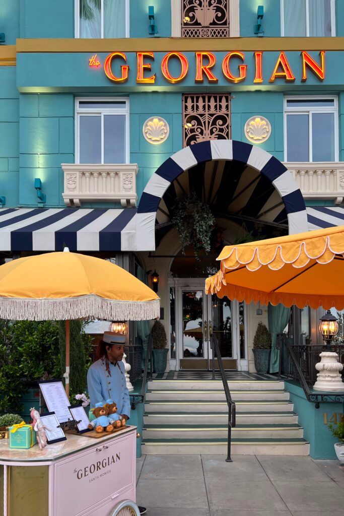 Exterior view of the entrance to the Georgian Hotel in Santa Monica with bellhop standing under yellow fringe umbrella.