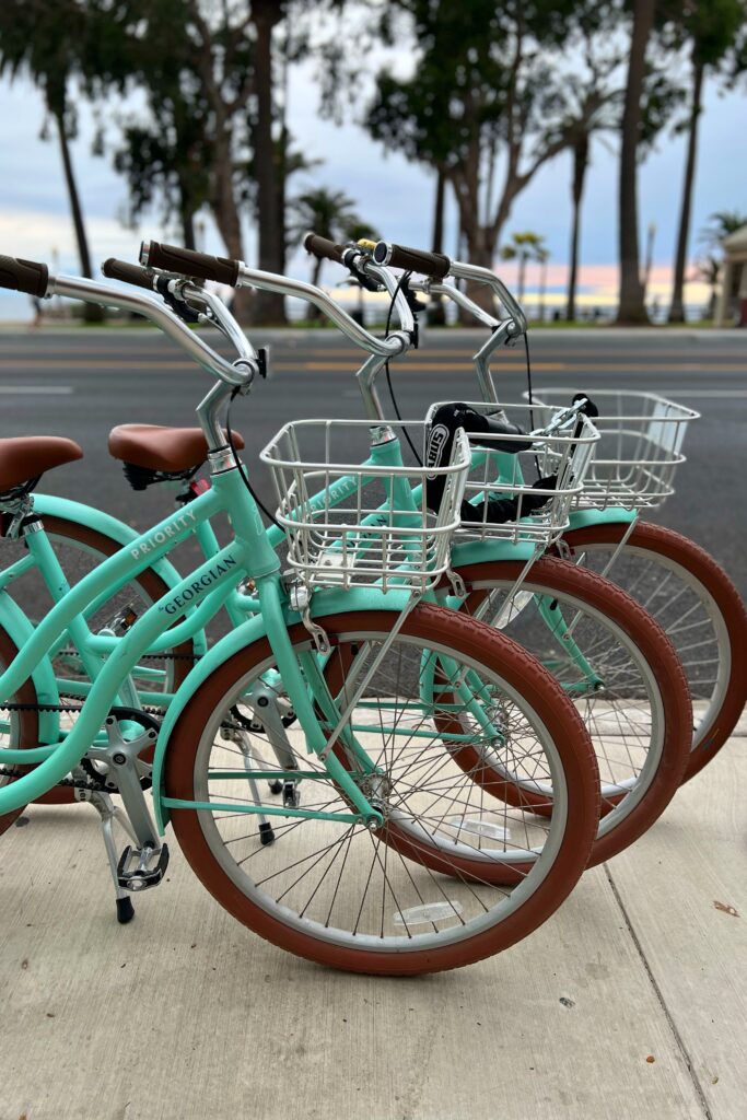 A row of charming turquoise bicycles with baskets with the Georgian Hotel in Santa Monica logo against the ocean.