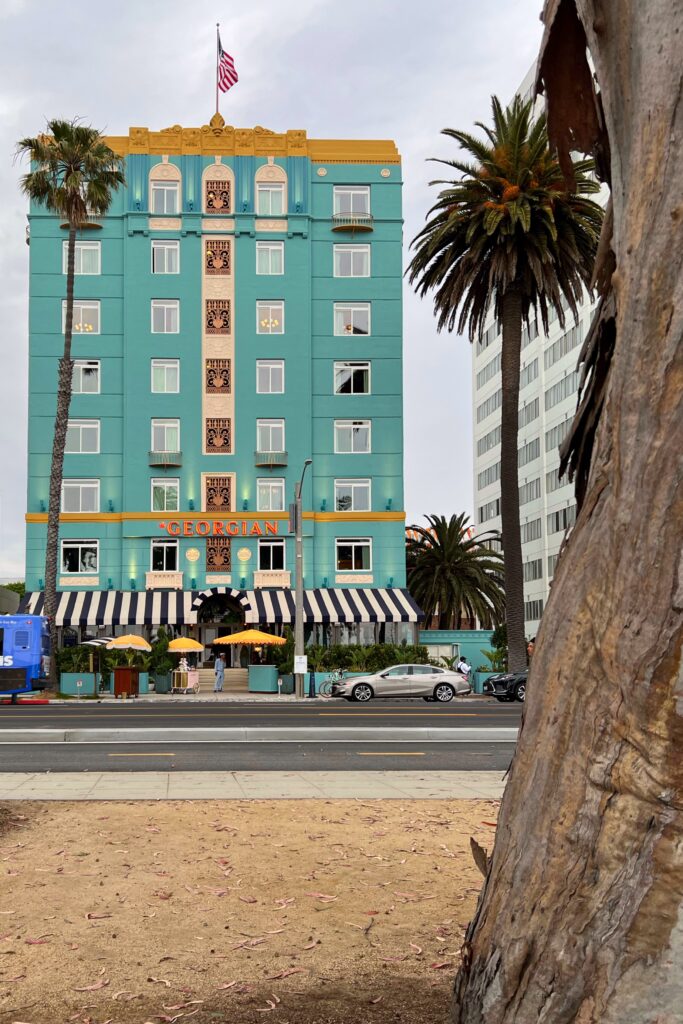 Exterior view of the turquoise Georgian Hotel in Santa Monica with striped awning and yellow fringe umbrellas with palm trees.
