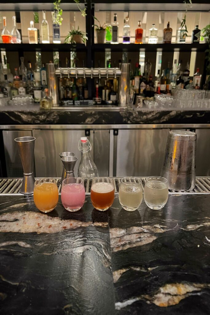 Mini flight of colorful craft cocktails lined up on a marble bar at 1 Hotel Nashville, with a backdrop of spirits and greenery.