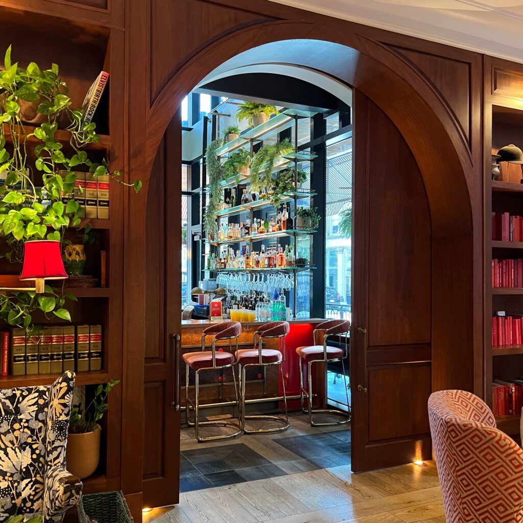 Inviting bar at Virgin Hotel New Orleans viewed through a wood archway, with pink bar stools, greenery, and floor-to-ceiling liquor shelves