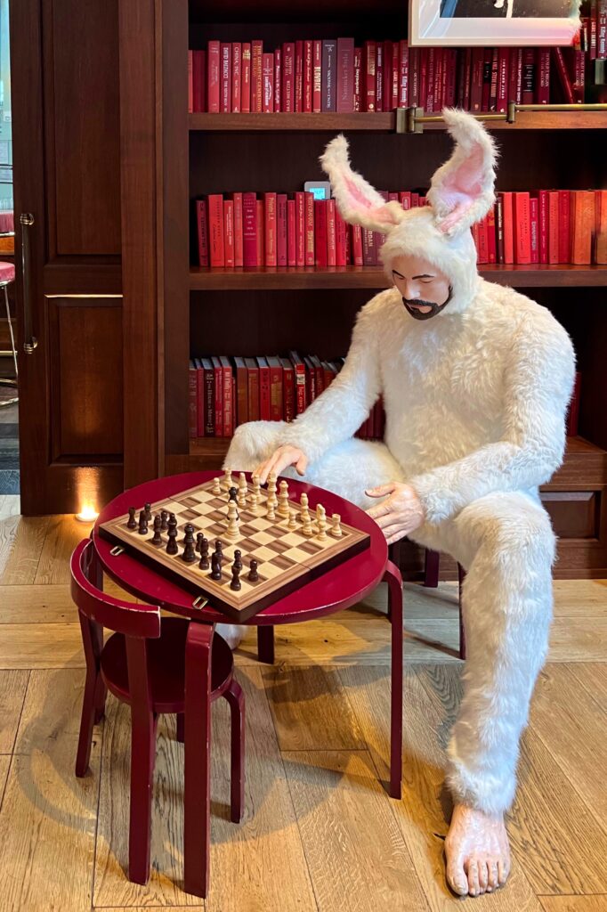 Eclectic seating including a whimsical rabbit sculpture playing chess in a library stacked with red books on the bookshelves.