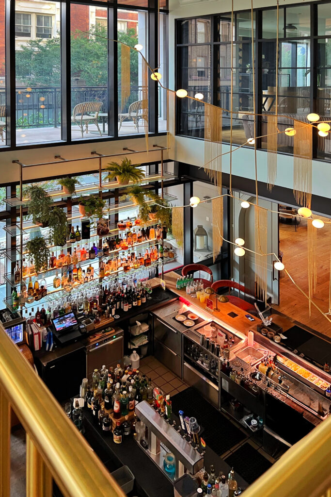 Overhead view of the bar at Virgin Hotel New Orleans with floor-to-ceiling windows, suspended glass shelving with plants and spirits, sculptural lighting installation, and open airy layout