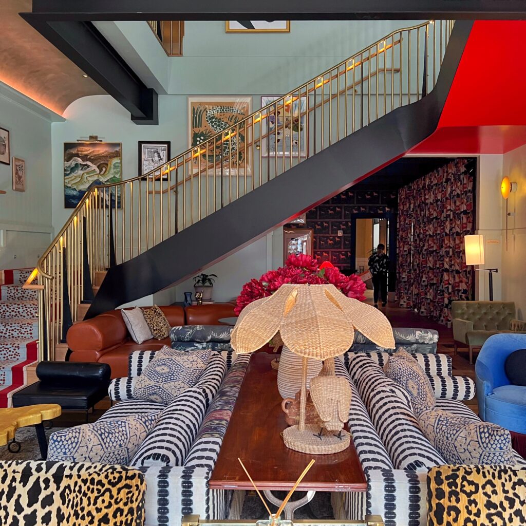 Vibrant lobby lounge at Virgin Hotel New Orleans featuring mix of animal print and geometric upholstered chairs, leather sectionals, rattan flower lamp, and striking gold stair railing