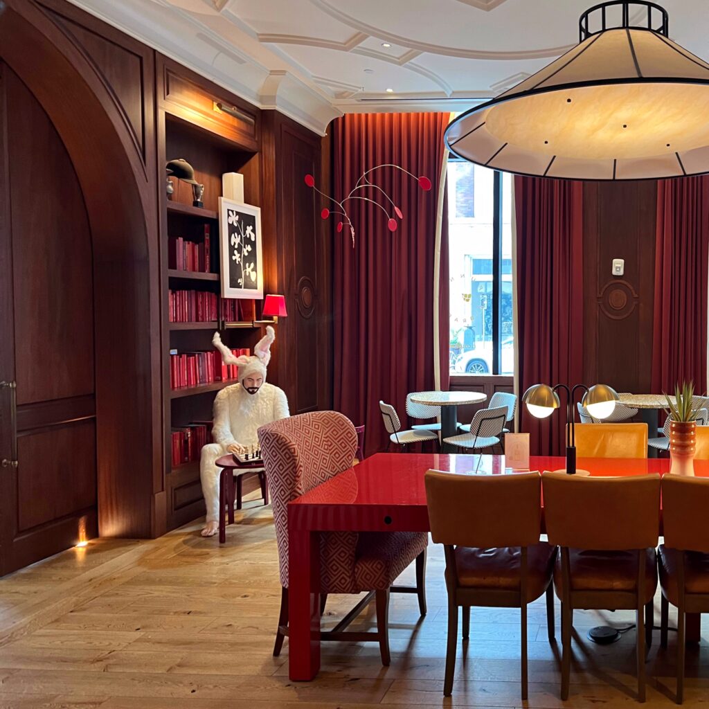 Eclectic hotel lounge at Virgin Hotels New Orleans with rich wood paneling, monochrome red bookshelves, statement lighting, and an unexpected rabbit-costumed figure at a chess table — a maximalist-meets-surrealist interior concept blending high design with humor.