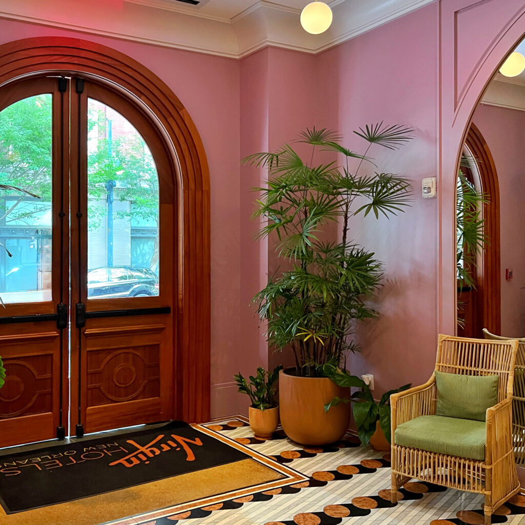 Boutique hotel entryway featuring blush pink walls, arched double doors, mosaic tile floor, rattan armchair, and potted greenery