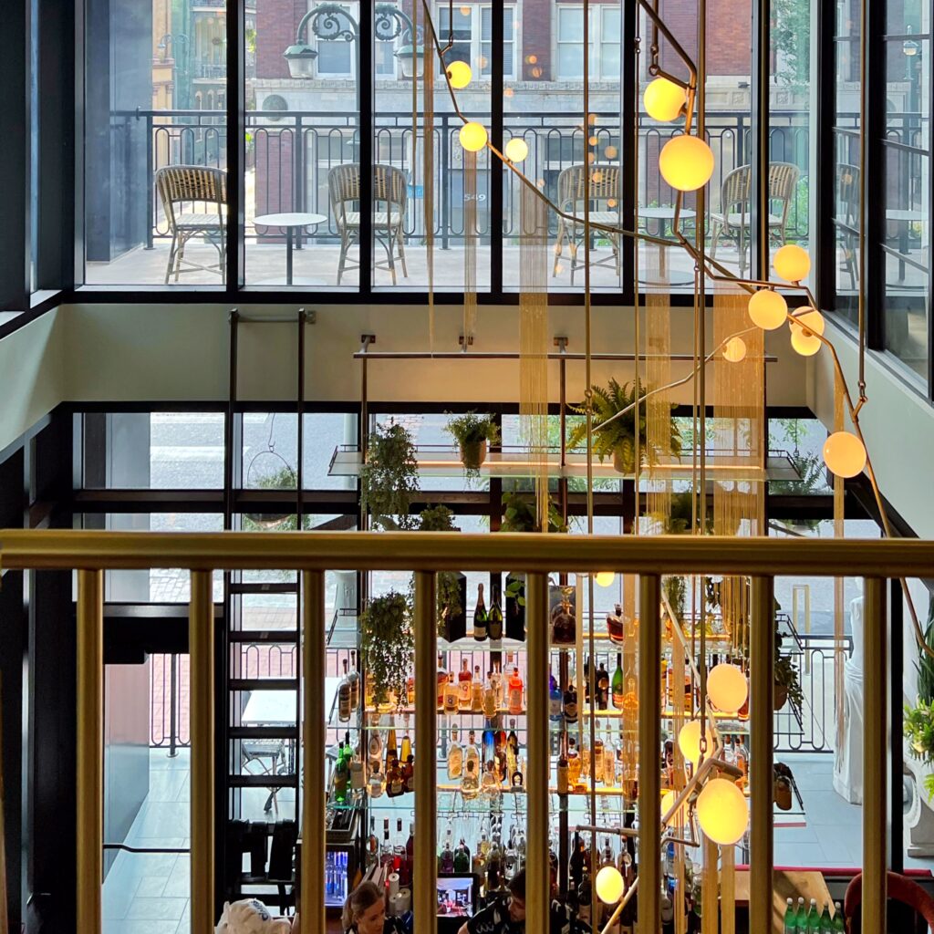 Modern bar at Virgin Hotel New Orleans with floor-to-ceiling windows, globe pendant lights, and a lush display of hanging plants