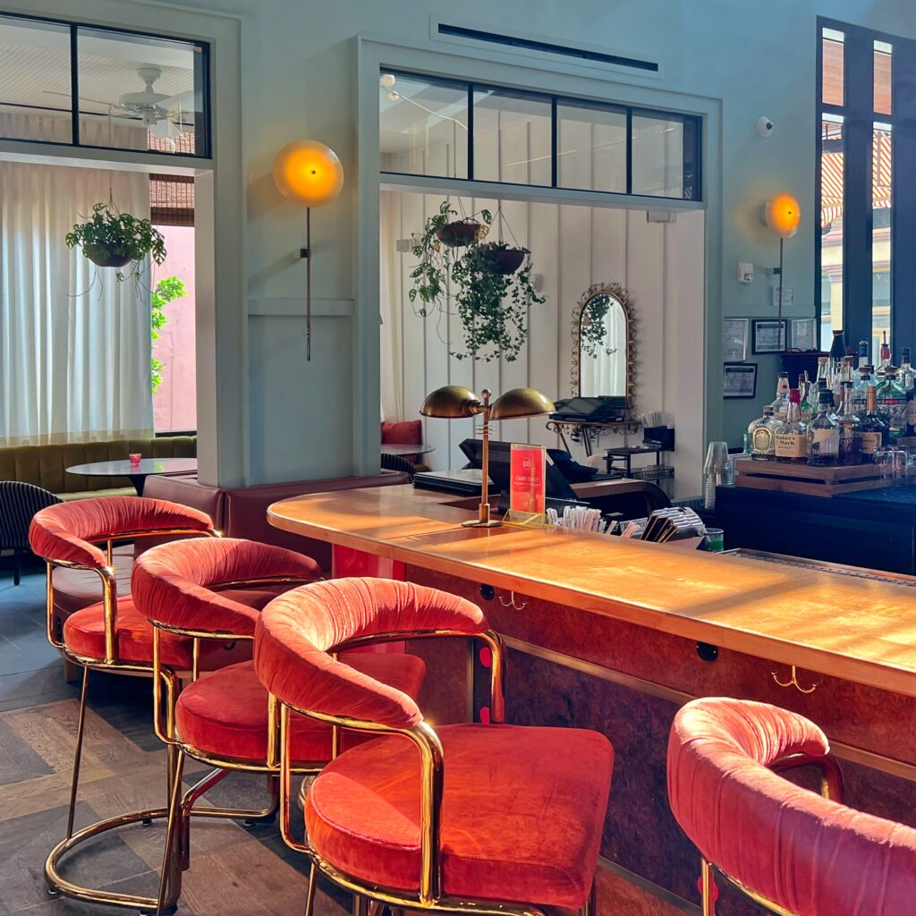 Sunny bar at Virgin Hotel New Orleans featuring curved brass-framed stools with red velvet seats, warm wood counter, and mid-century inspired lighting