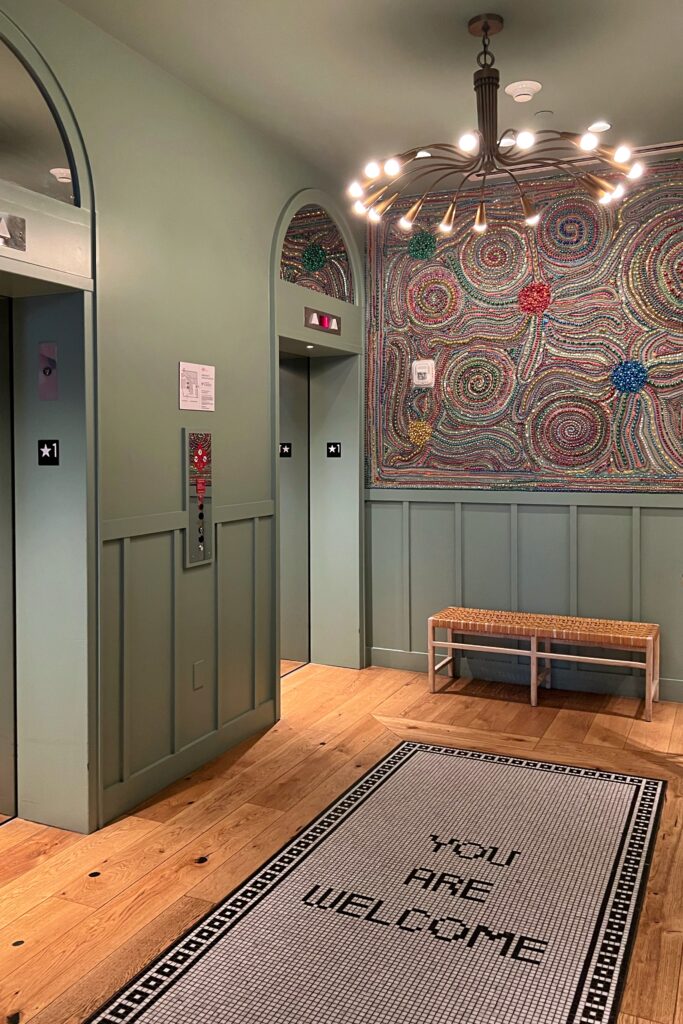 Colorful elevator lobby at Virgin Hotel New Orleans with mosaic wall art, sage green paneling, and a tile rug reading “You Are Welcome”