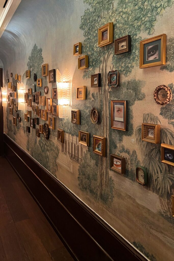 Art-filled corridor inside Cafe Carmellini, featuring a dramatic gallery wall of miniature gilded frames displaying surreal eye illustrations set against hand-painted scenic wallpaper. Soft sculptural wall sconces illuminate the hallway, blending Old World European salon style with contemporary hospitality design in one of Manhattan’s most visually distinctive restaurant interiors.
