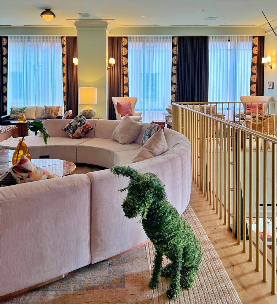 Playful hotel lounge at Virgin Hotel New Orleans, featuring a curved upholstered sofa, geometric-patterned throw pillows, amber glass vase centerpiece, gold railing, and whimsical green topiary dog accent