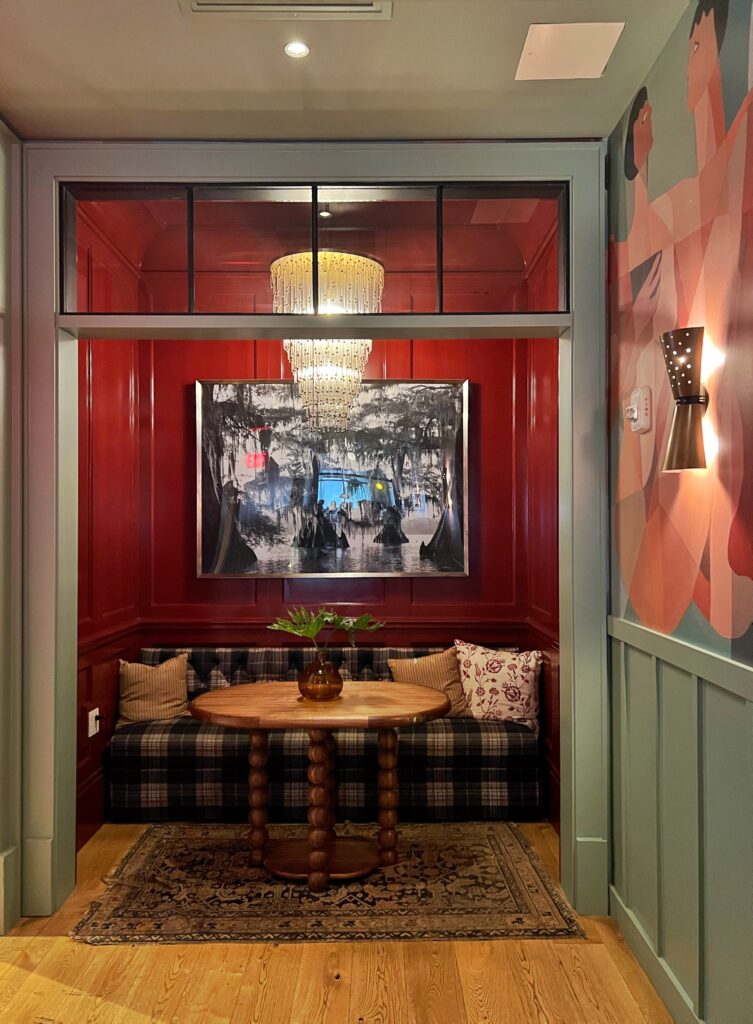 Intimate dining nook at Virgin Hotel New Orleans, Cozy alcove with glossy red paneled walls, plaid upholstered bench, sculptural wood table, amber vase centerpiece, layered rug, and tiered crystal chandelier