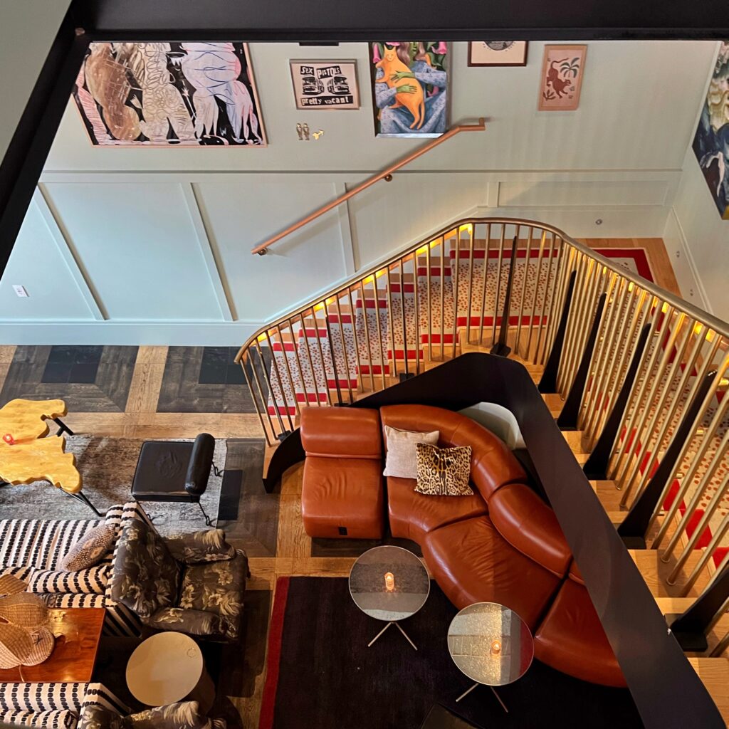 Overhead view looking into lobby at Virgin Hotel, New Orleans: featuring a caramel-brown curved leather sectional, gold balustrade staircase with red-and-white patterned runner, black-and-white upholstered armchairs, eclectic art pieces, and a mix of wood and glass tables on a patterned floor