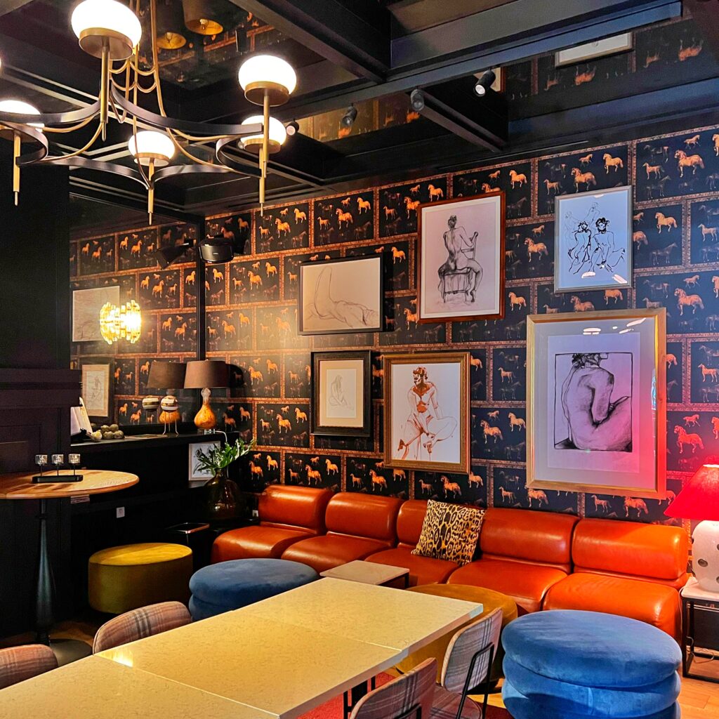 Eclectic lounge at Virgin Hotel New Orleans with cognac leather banquette, velvet stools, patterned wallpaper with dog motifs, framed figure sketches, modern chandelier, and layered warm-toned lighting