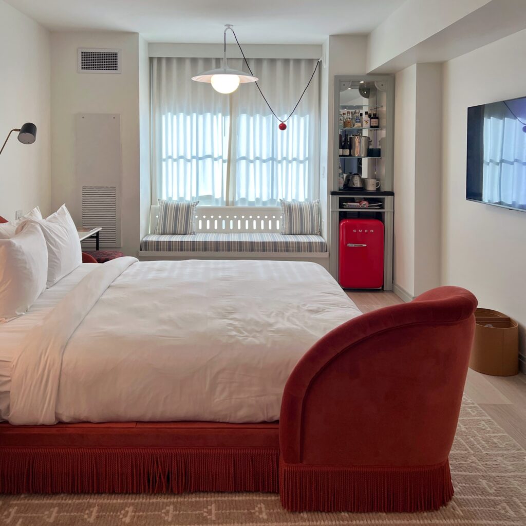 Guest room at Virgin Hotel New Orleans eaturing curved red velvet bed with fringe detail, built-in window bench, striped upholstery, and retro-style red Smeg refrigerator