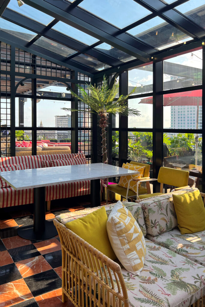 Rooftop lounge at Virgin Hotel New Orleans featuring marble-topped table, wicker sofa with fern print cushions, mustard yellow pillows, and black grid-frame windows