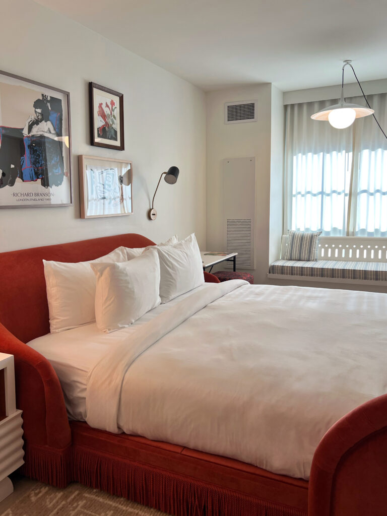 Guest room at Virgin Hotel New Orlean featuring curved red velvet bed with fringe detail, white linens, wall-mounted reading light, and framed art