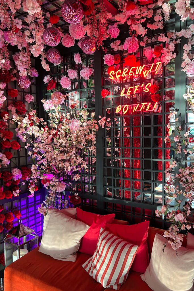 Virgin Hotel New Orleans rooftop lounge corner with hanging pink blossoms, mirrored lattice wall, red and white striped pillows, and neon sign reading “Screw It Let’s Do It"