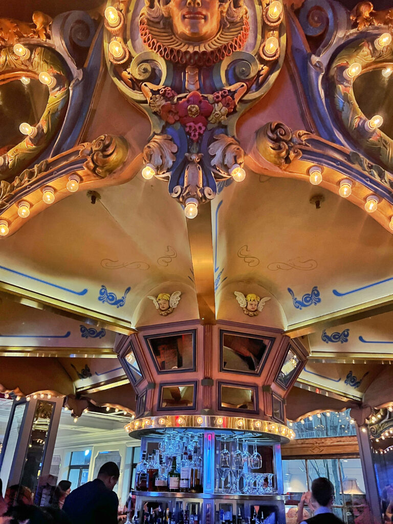 The iconic Carousel Bar in New Orleans, Close-up of a carousel-style ceiling with ornate painted faces, floral details, golden lighting, and mirrored panels above a rotating circular bar stocked with bottles and glassware