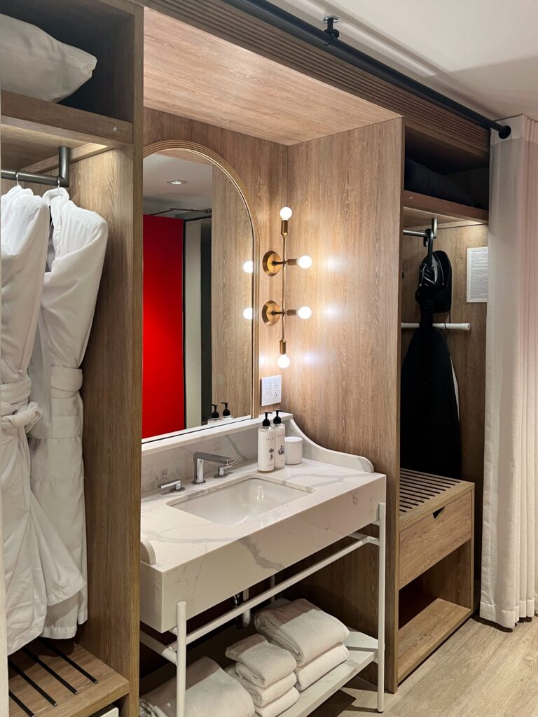 Guest room vanity at Virgin Hotel New Orleans featuring arched mirror with globe lights, white marble sink, wood cabinetry, and open wardrobe storage