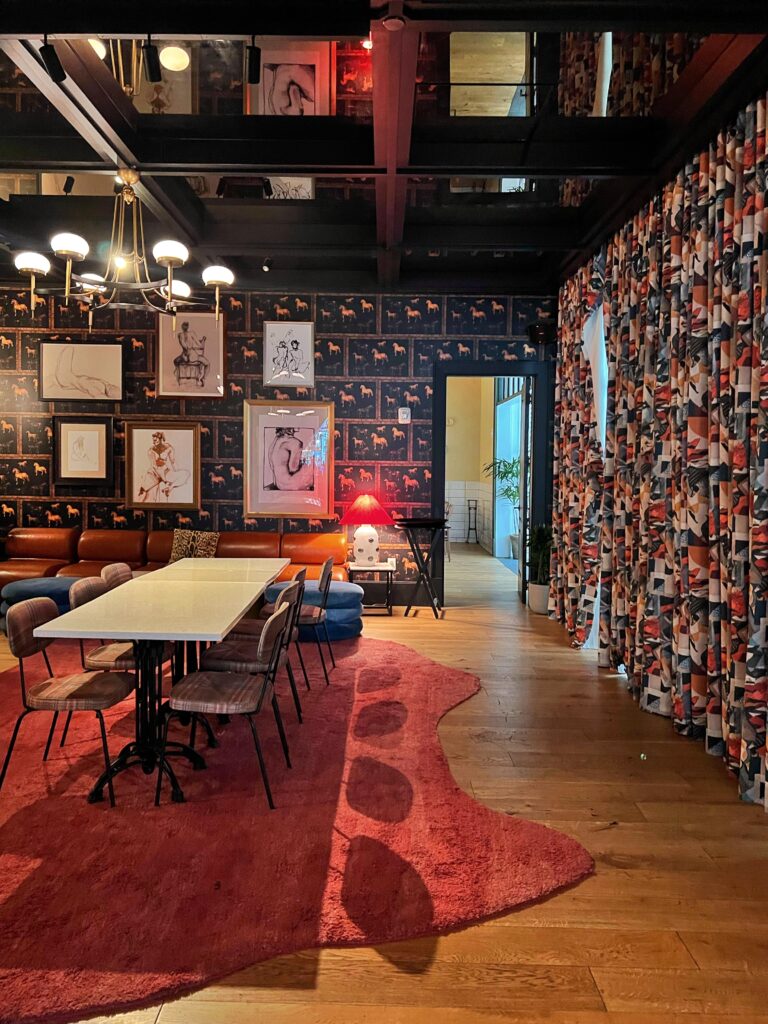 Eclectic lounge at Virgin Hotel New Orleans, Playful interior featuring a red abstract rug, plaid-upholstered dining chairs, marble-top communal table, and gallery wall with line art and matisse-themed wallpaper