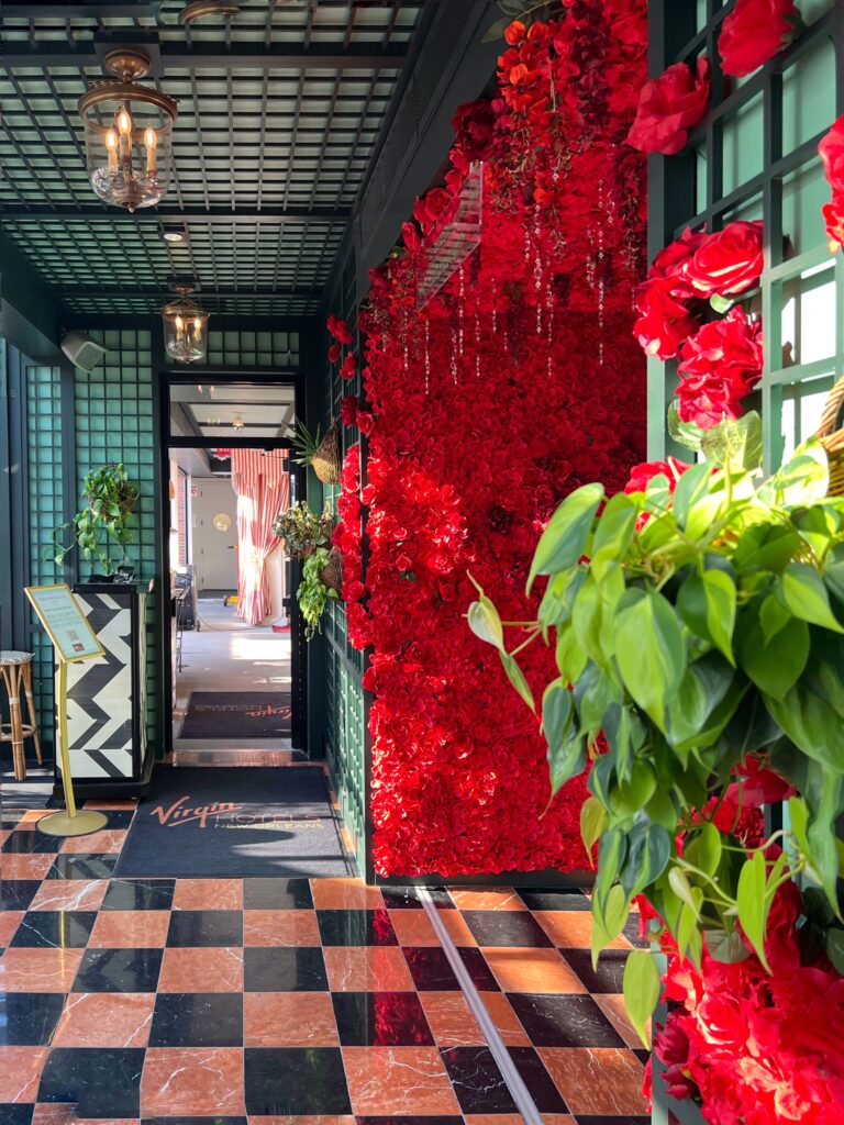 Vibrant entrance to Rooftop at Virgin Hotel New Orleans featuring lush red floral installation, black and pink marble checkerboard flooring, and grid-patterned green ceiling