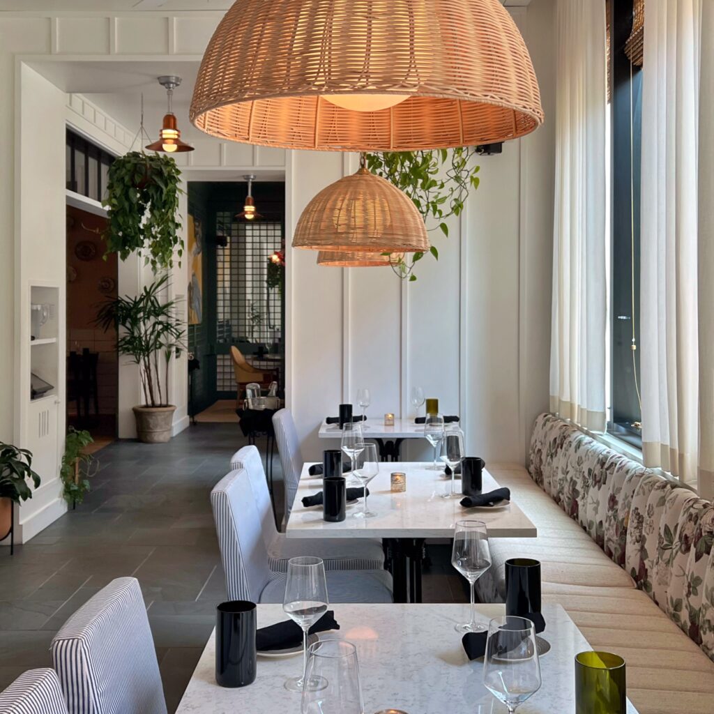 Light-filled dining at the Commons Club restaurant in the Virgin Hotel, New Orleans featuring woven rattan dome pendant lights, black glassware, soft neutral seating, and hanging plants.
