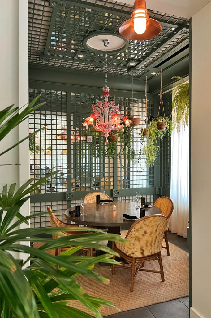Cozy private dining nook inside the Commons Club restaurant in the Virgin Hotel, New Orleans ramed by green lattice walls, decorated with hanging greenery and an ornate pink Murano-style glass chandelier above a dark wood table