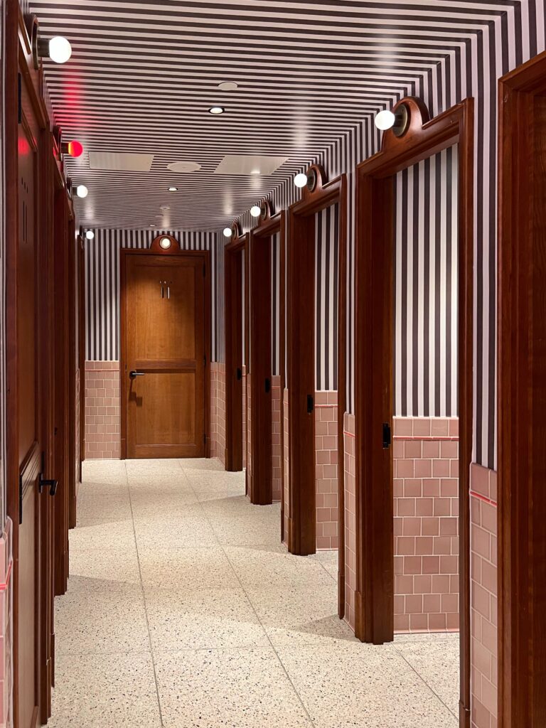 Bold restroom interior at Virgin Hotel New Orleans with continuous vertical black and white stripes across walls and ceiling, pink subway tile wainscoting, terrazzo floor, and globe sconces above wood-framed doorways