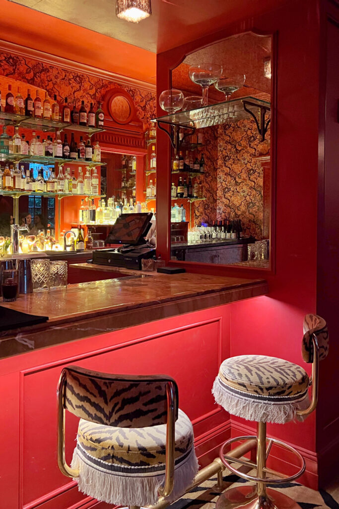 Intimate view of Bar Marilou in New Orleans, Luxurious red-walled bar with ornate patterned wallpaper, marble countertop, mirrored shelves lined with liquor bottles, and two gold-framed bar stools with tiger-print upholstery and fringe trim