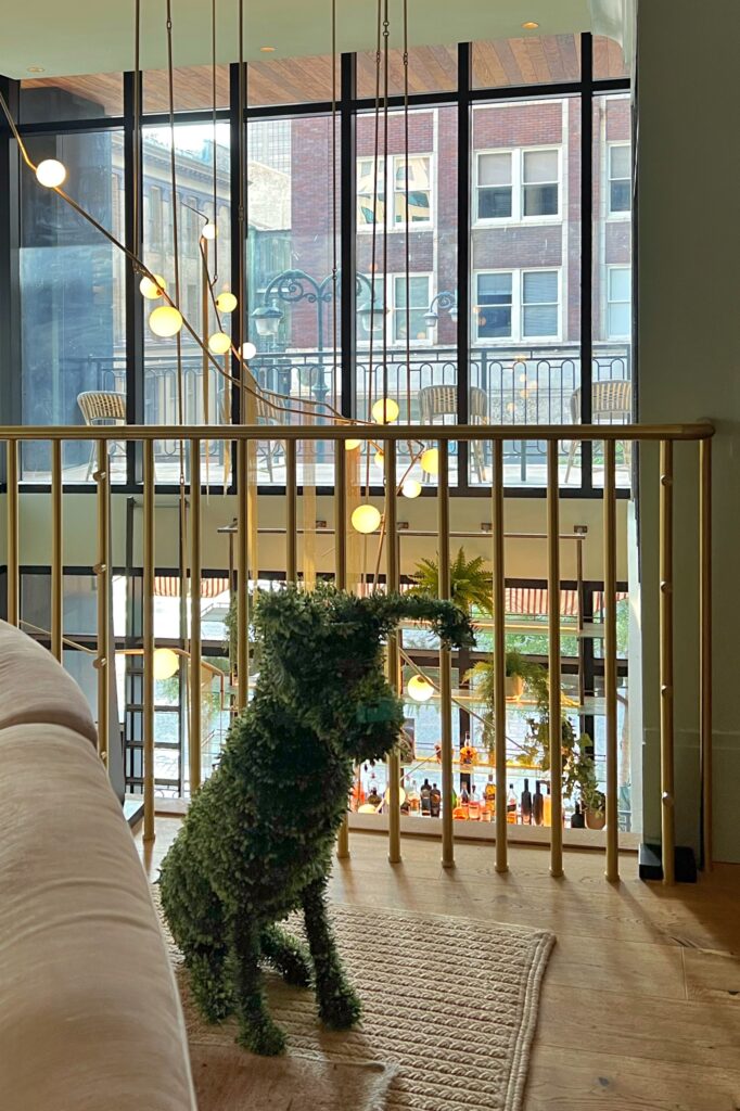 Playful topiary dog sculpture in the upstairs lounge of Virgin Hotel New Orleans, Greenery-covered dog-shaped sculpture placed near gold railing in a modern loft space with floor-to-ceiling windows and globe pendant lights