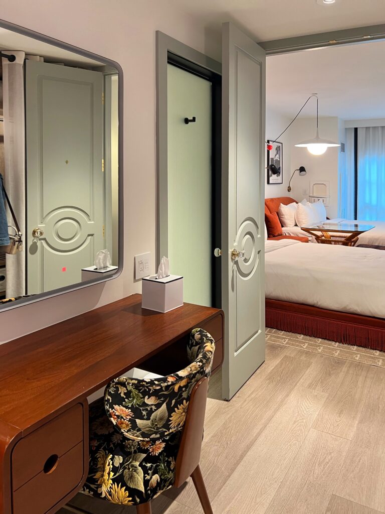 Guest room vanity area at Virgin Hotel New Orleans featuring mid-century style wood vanity, floral patterned chair, paneled doors, and glimpse of bedroom with fringe-trimmed red velvet bed