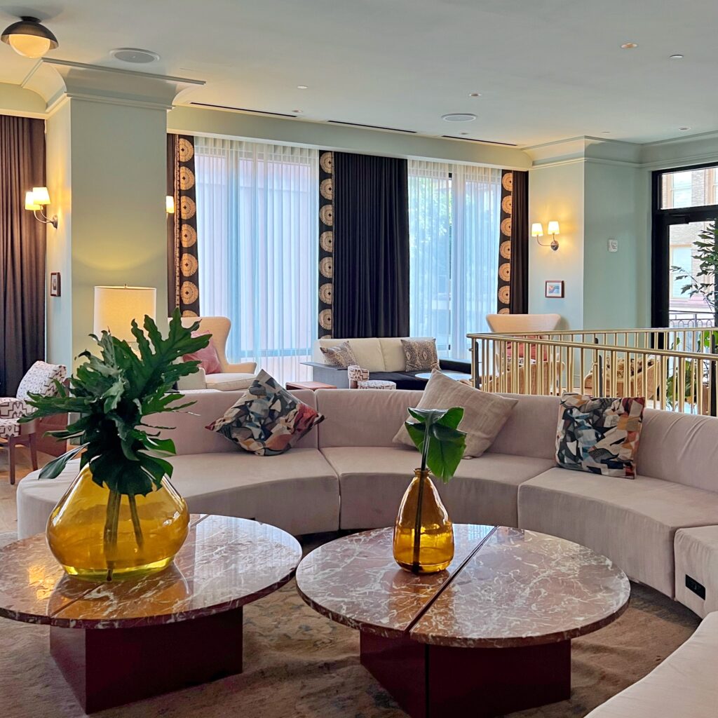 Chic upstairs lounge at Virgin Hotel New Orleans, Elegant seating area featuring curved modular sofas, geometric-patterned throw pillows, round red marble coffee tables, and floor-to-ceiling windows framed by dark drapes with gold medallion trim