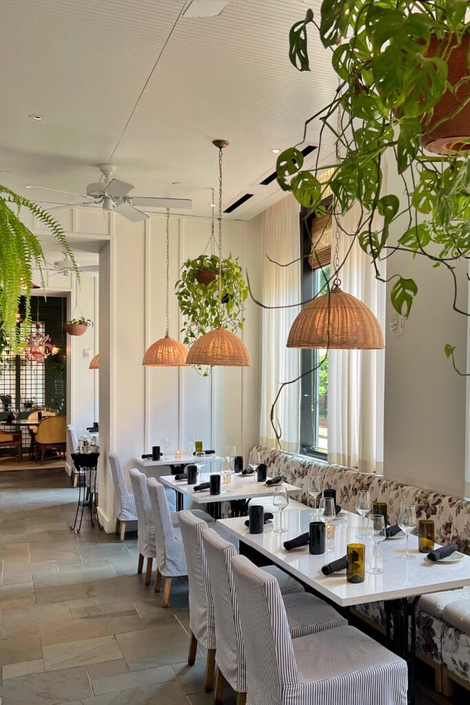 Airy dining space at Virgin Hotel New Orleans Light-filled restaurant interior featuring marble tables, striped slipcovered chairs, floral banquette seating, and natural rattan light fixtures with hanging plants.