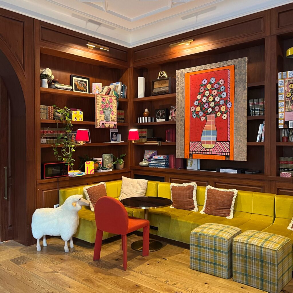 Colorful lobby reading nook at Virgin Hotel New Orleans featuring a mustard velvet banquette, red accent chair, plaid cube stools, whimsical sheep sculpture, and vibrant floral artwork