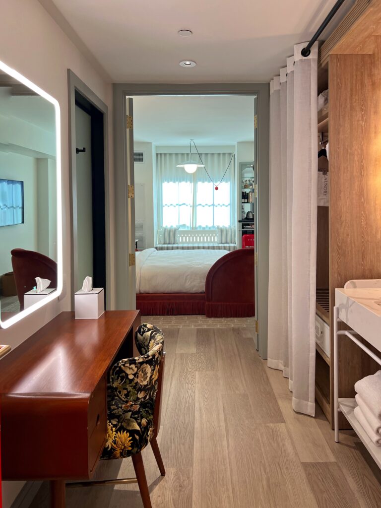 Guest room at Virgin Hotel New Orleans featuring wood vanity with floral upholstered chair, light wood floors, and bedroom with curved red velvet bed
