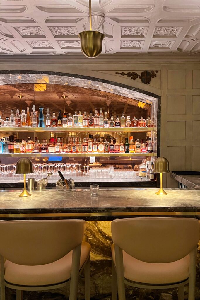 Bar at Drusie & Darr inside the Hermitage Hotel in Nashville, with a polished marble counter, brass table lamps, cream upholstered bar chairs, and a backlit stone arch framing shelves of spirits beneath a carved coffered ceiling — a refined southern bar interior design.