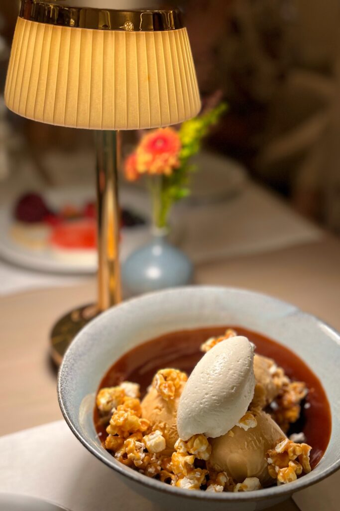Dessert at Drusie & Darr inside the Hermitage Hotel in Nashville, featuring scoops of caramel ice cream with caramel popcorn and vanilla cream served in a ceramic bowl, styled beneath a brass table lamp with modern dining accents — blending culinary creativity with refined restaurant design.