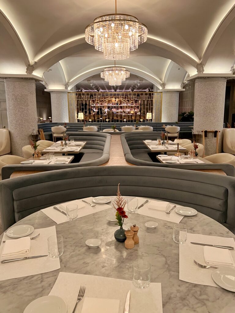 Dining room at Drusie & Darr inside the Hermitage Hotel in Nashville, with vaulted ceilings, sparkling crystal chandeliers, curved velvet banquettes, marble dining tables, and a brass screen framing the view toward the marble bar — a refined example of contemporary restaurant interior design in the South.