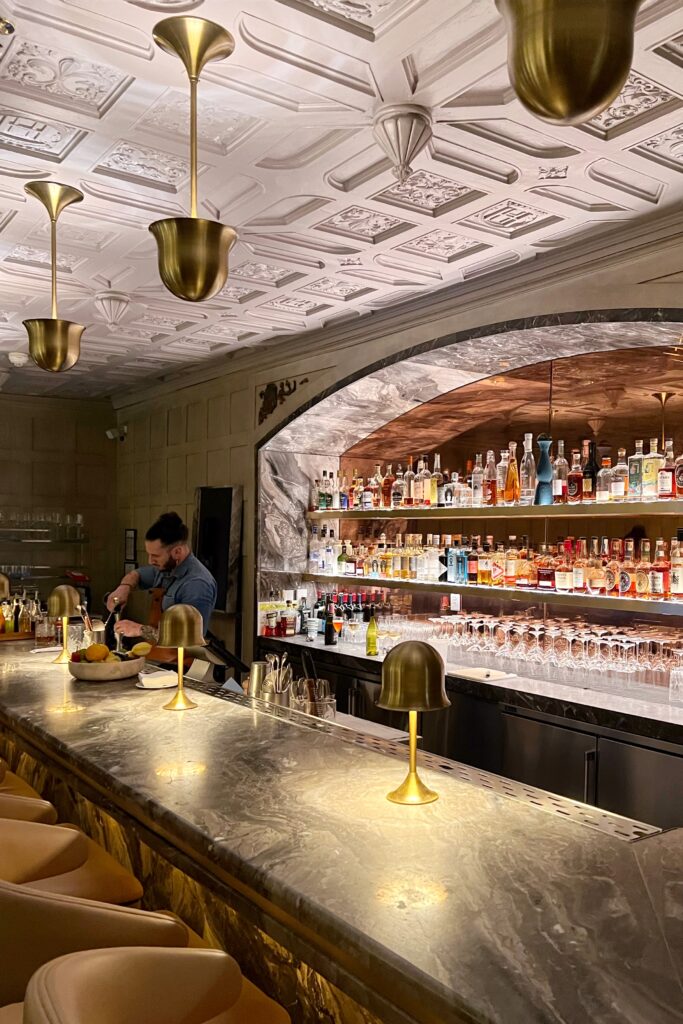 Marble bar at Drusie & Darr inside the Hermitage Hotel in Nashville, with brass pendant lighting, a carved coffered ceiling, and a backlit stone arch showcasing rows of spirits — a standout example of Nashville bar interior design.